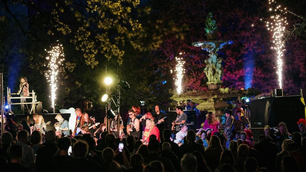 An ensemble of guitarists are well lit, as audience looks on and sparks illuminate the staging