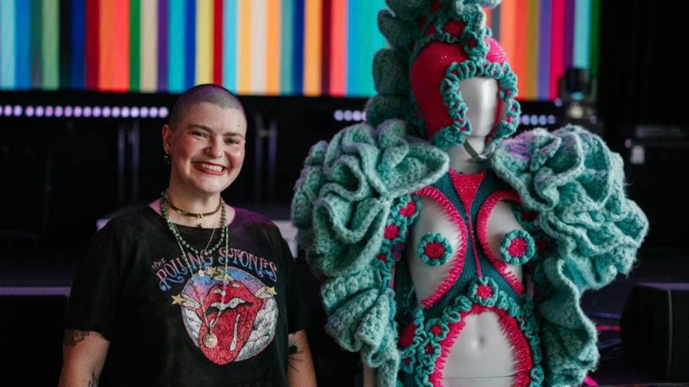 A woman standing next to handmade wearable yarn art.