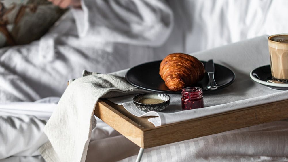 A person sitting in the bed with a tray with a croissant and a coffee